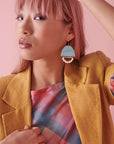 A lady with pink hair sits against a pink background and models a sideview of the Lovelace earrings in sky. She has one arm raised resting on her head, the other hand holds the lapel of her mustard jacket. She wears a pink and blue check top under the jacket.