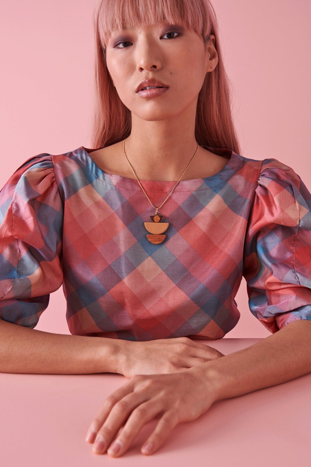 A lady with pink hair sits against a pink background. She models the Marcel necklace in mustard and wears a pink and blue checkered top.