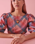 A lady with pink hair sits against a pink background. She models the Marcel necklace in mustard and wears a pink and blue checkered top.