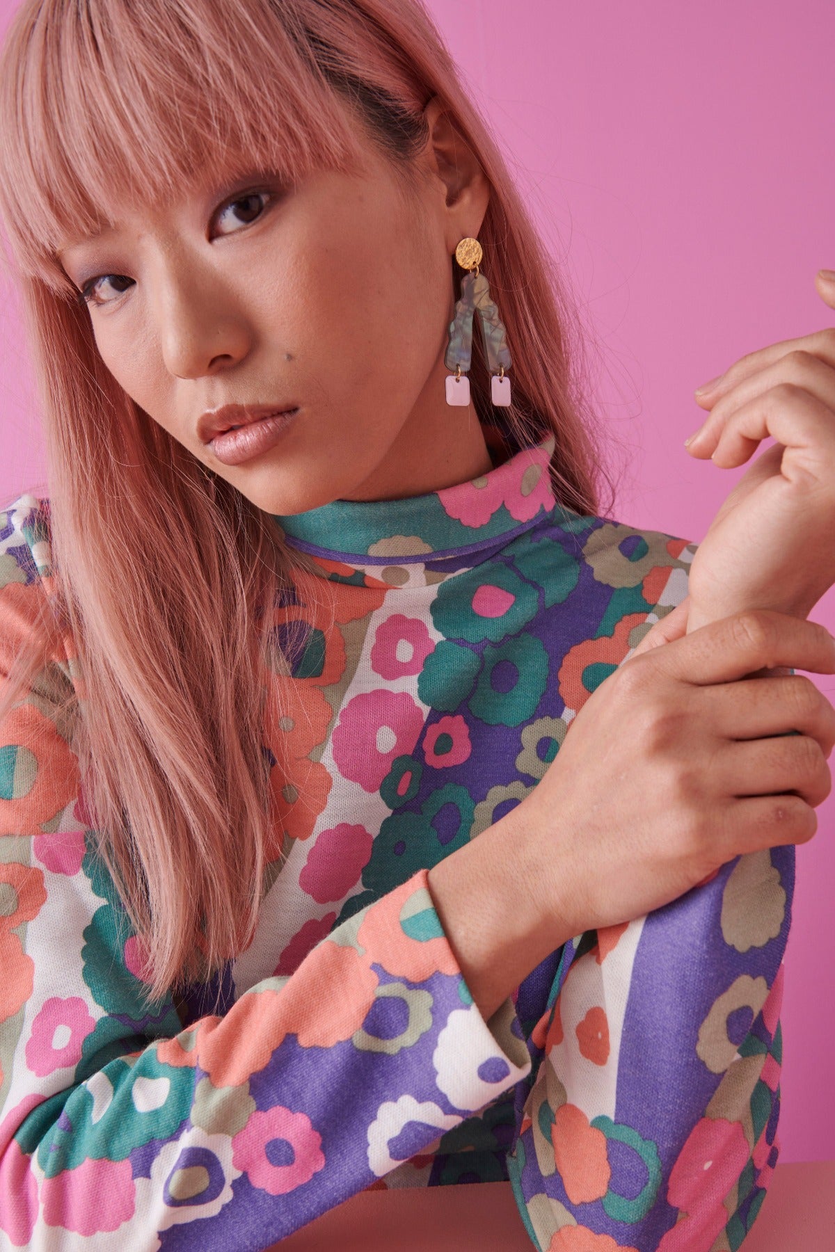 A lady with pink hair sits against a bright pink background and models a sideview of the Groove Thang earrings in green. She holds her wrist in one hand and wears a multicoloured retro floral top.