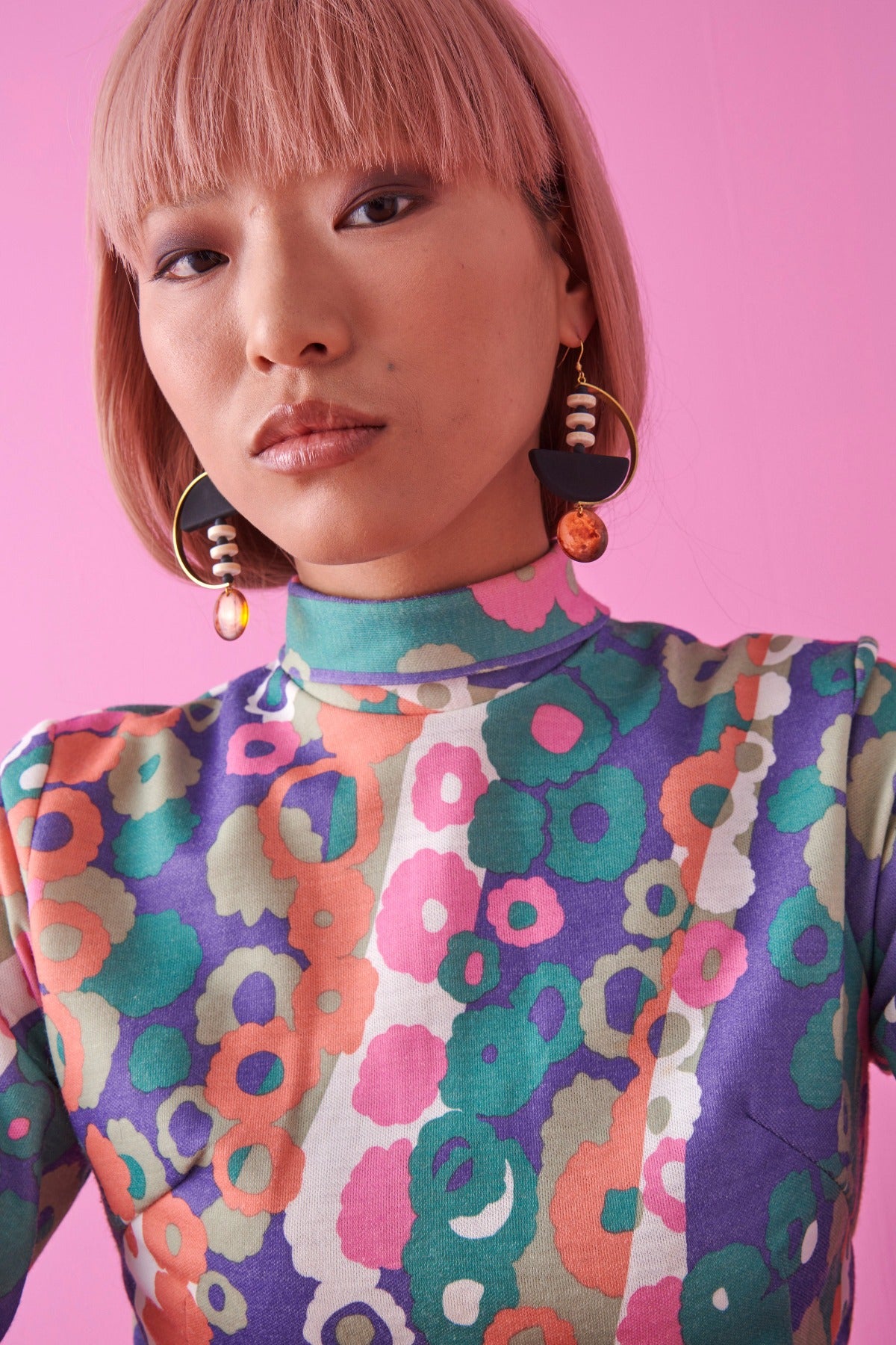 A lady with pink hair models the Monument earrings in black. She sits against a pink background and wears a multicoloured retro floral print top.