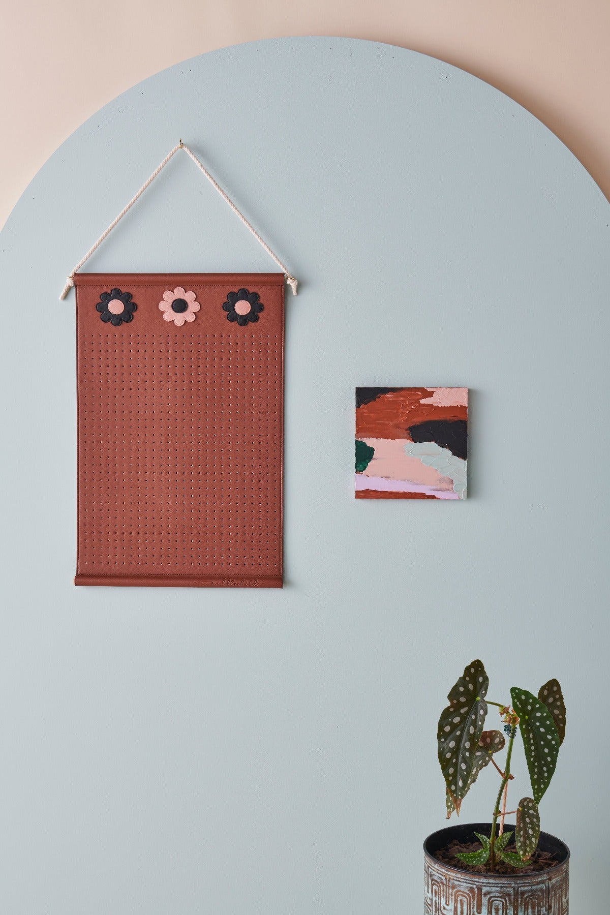 A brown wall hanger with three brown and pink flowers at the top is hung by a rope and styled against a blue arch and pink wall. Next to the wall hanger is a small square abstract painting and a pot plant with spotted leaves sits below the painting.