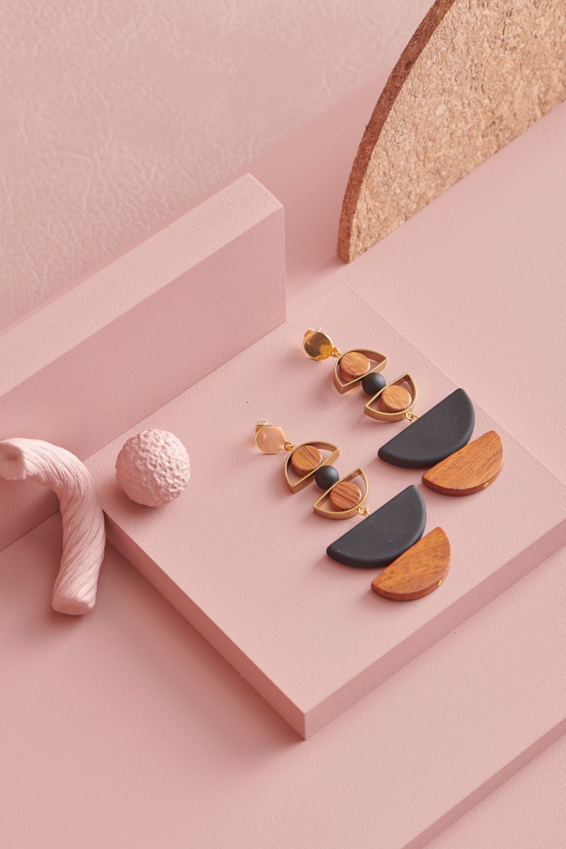 A pair of Marcel earrings in black sit styled against a pink background, with a cork arch detail and textured pink elements in frame.