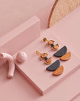 A pair of Marcel earrings in black sit styled against a pink background, with a cork arch detail and textured pink elements in frame.
