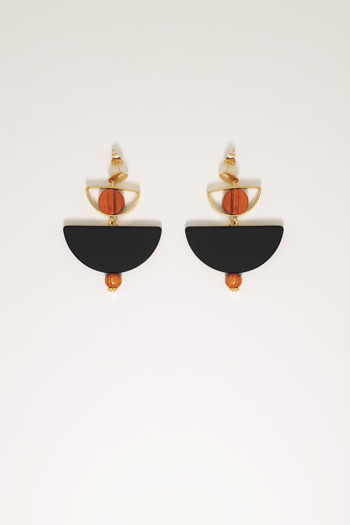 A pair of stud dangle earrings sit against a white background. They feature a  small half circle brass piece with a wooden circle bead enclosed, below this hangs a black D shape bead followed by a small wooden bead and a tiny gold bead.
