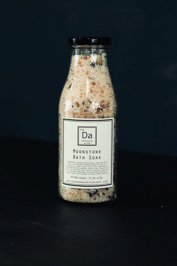 A glass jar filled with bath crytstals and petals sits against a black background. It has a white label with a black border and inside the text Moonstone Bath Soak is written along with a list of ingredients.
