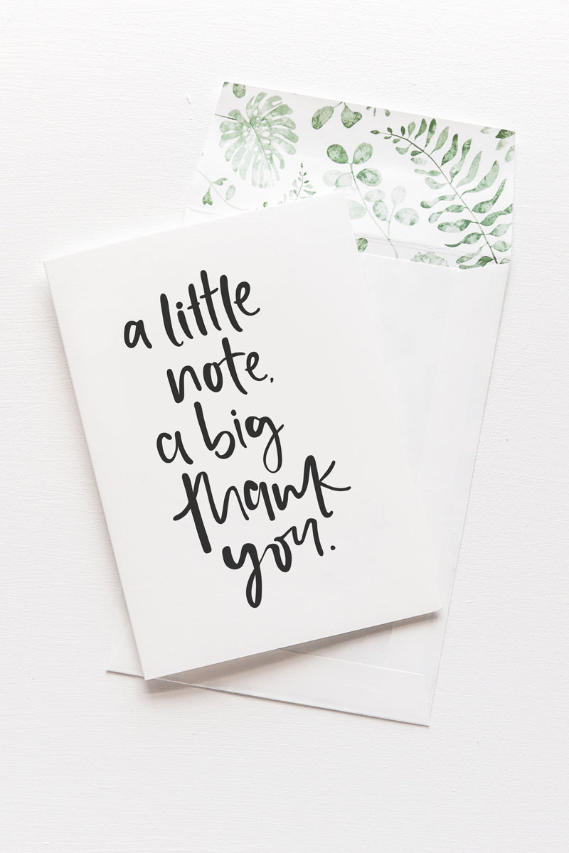 A white card with the message 'a little note. a big thankyou.' is sitting on a white background. The card uses black script font. There is a white envelope behind the card which has a green watercolour plant design inside.