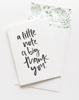 A white card with the message 'a little note. a big thankyou.' is sitting on a white background. The card uses black script font. There is a white envelope behind the card which has a green watercolour plant design inside.