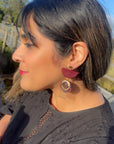 A lady with dark hair and red lipstick sits outside. She models a sideview of the bordo red Broadcast earrings and wears a black top.