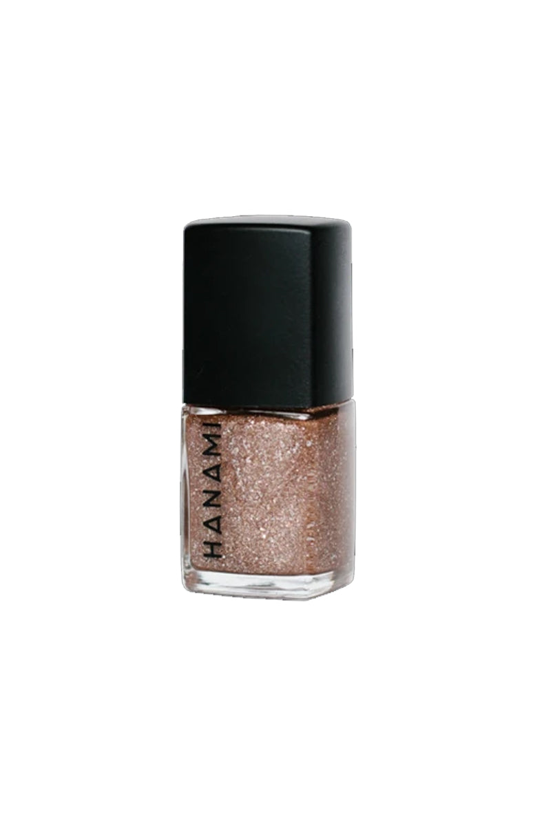 A bottle of nail polish in sits against a white background. It features a cubed glass bottle with the words HANAMI written in black uppercase text along one side and has a black cube-shaped lid. It is filled with a rose gold glitter nail polish.