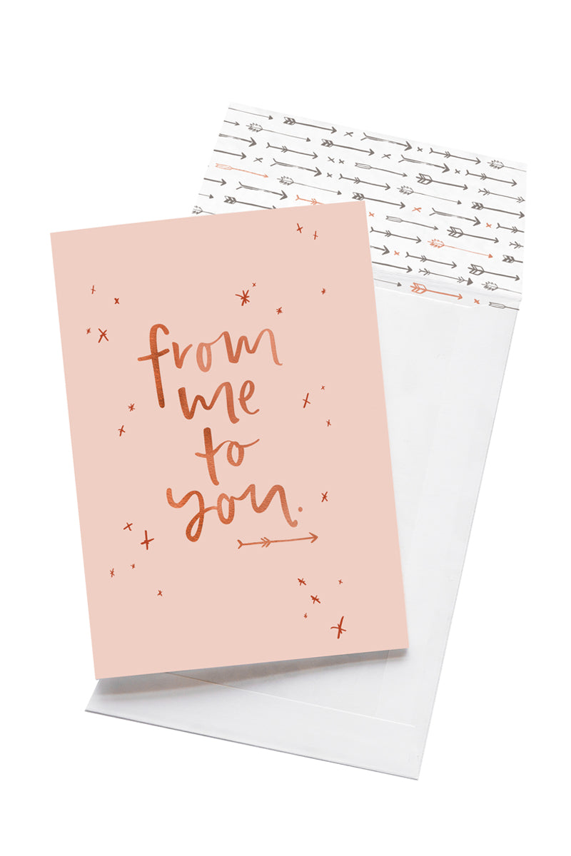 A pale pink card with the message 'from me to you' is sitting on a white background. The card uses rose gold script font and has hand drawn stars around it. There is a white envelope behind the card which has an arrow pattern design inside.