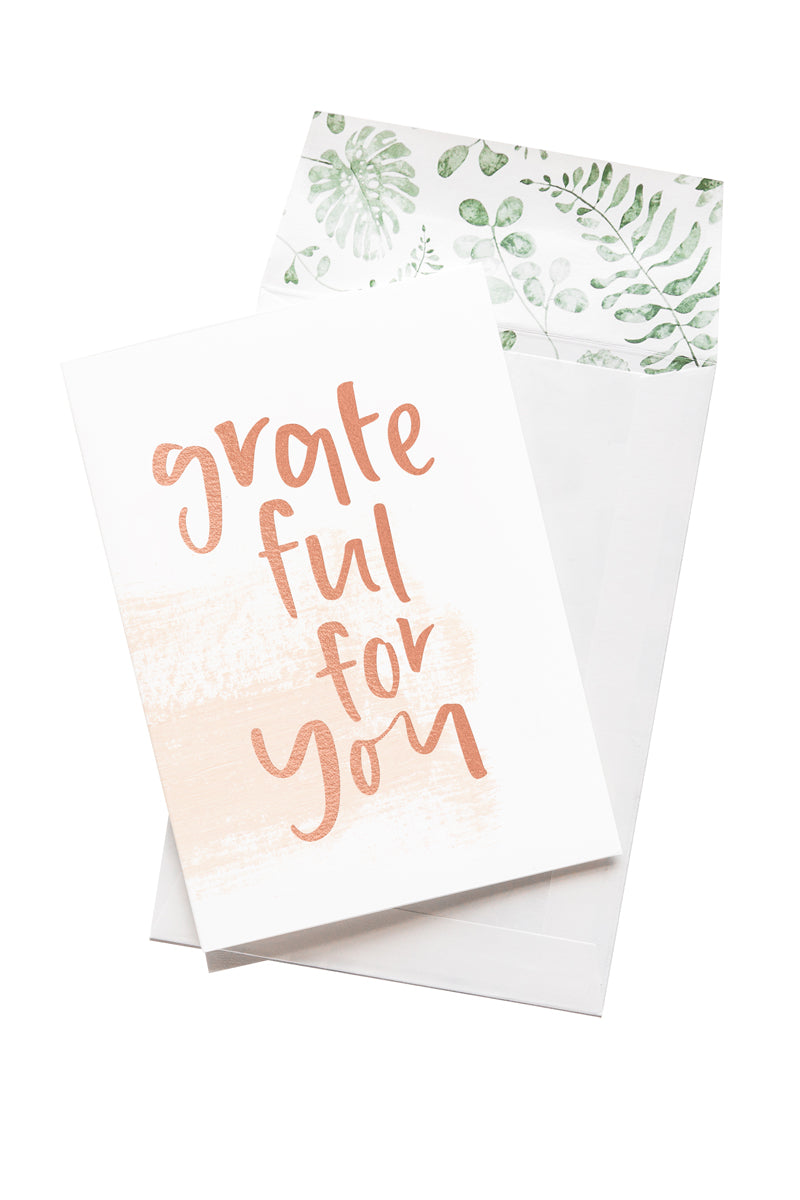 A white card with the message 'grateful for you' is sitting on a white background. The card uses rose gold script font. There is a white envelope behind the card which has a green watercolour plant design inside.