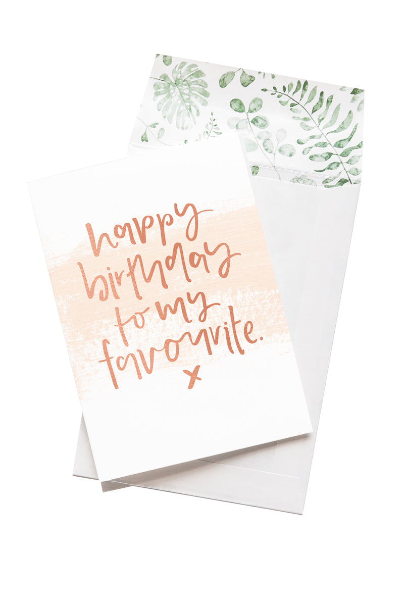 A white card with the message 'happy birthday to my favourite. x' is sitting on a white background. The card uses rose gold script font and has a pale pink brush stroke behind the message. There is a white envelope behind the card which has a green watercolour plant design inside.