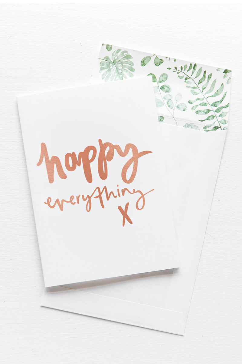 A white card with the message 'happy everything x' is sitting on a white background. The card uses rose gold script font. There is a white envelope behind the card which has a green watercolour plant design inside.