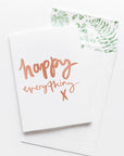A white card with the message 'happy everything x' is sitting on a white background. The card uses rose gold script font. There is a white envelope behind the card which has a green watercolour plant design inside.