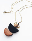 A necklace with a gold chain sits against a white background. It features a small wooden bead, followed by a brass D shape with a wooden circle bead enclosed, followed by a black D shaped bead, and a wooden D shape bead.