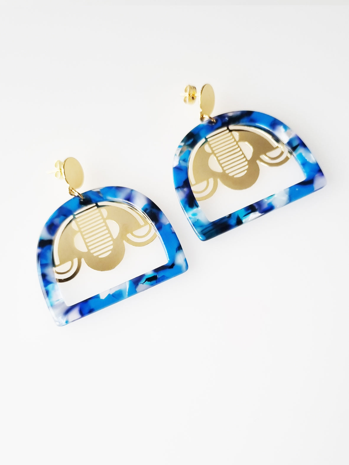 A pair of stud dangle earrings sits against a white background. They feature a blue multi-coloured acrylic arch shape and a custom art deco brass piece.