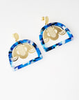 A pair of stud dangle earrings sits against a white background. They feature a blue multi-coloured acrylic arch shape and a custom art deco brass piece.