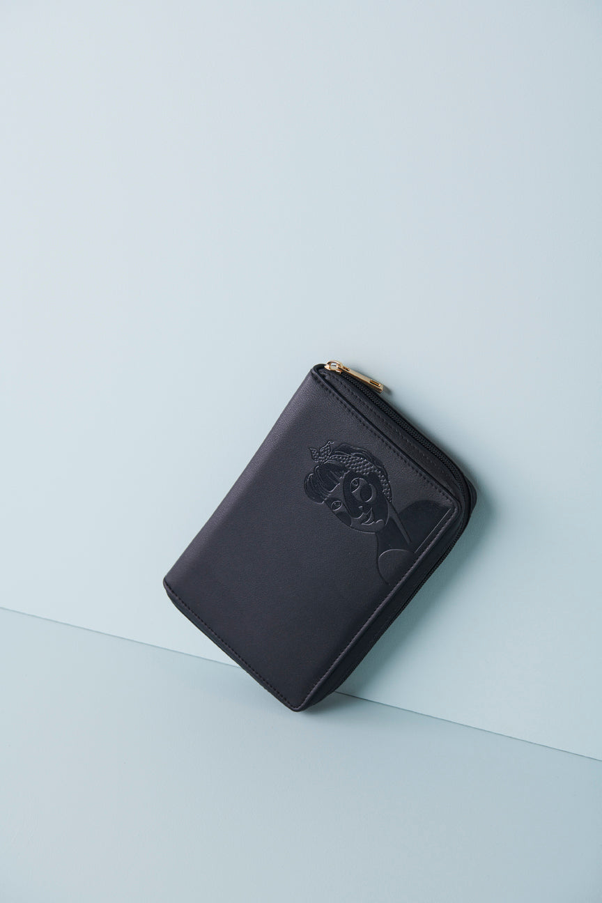 A black jewellery wallet sits on an angle against a baby blue background. The wallet features an embossed image of a lady with a fringe and head scarf. It has a black zip and has a gold zip pull.