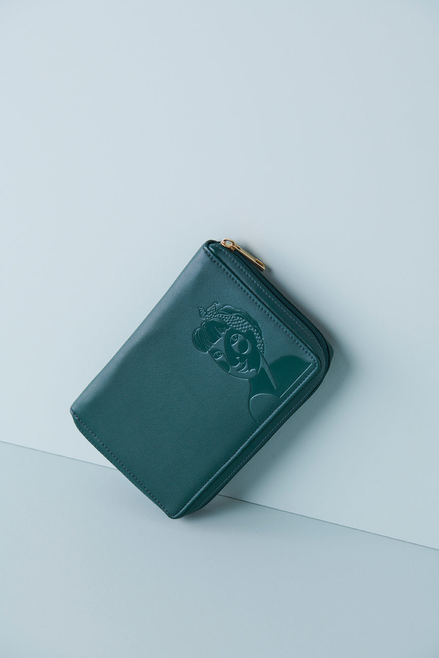 A forest green jewellery wallet sits on an angle against a baby blue background. The wallet features an embossed image of a lady with a fringe and head scarf. It has a green zip and has a gold zip pull.