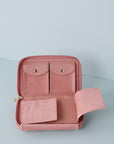 A pink wallet sits open against a baby blue background. The wallet inner is pink, the top section featues two felt pockets with pu flaps and gold clasp buttons. The bottom section features felt flaps that open to reveal a perforated pu section for earrings.