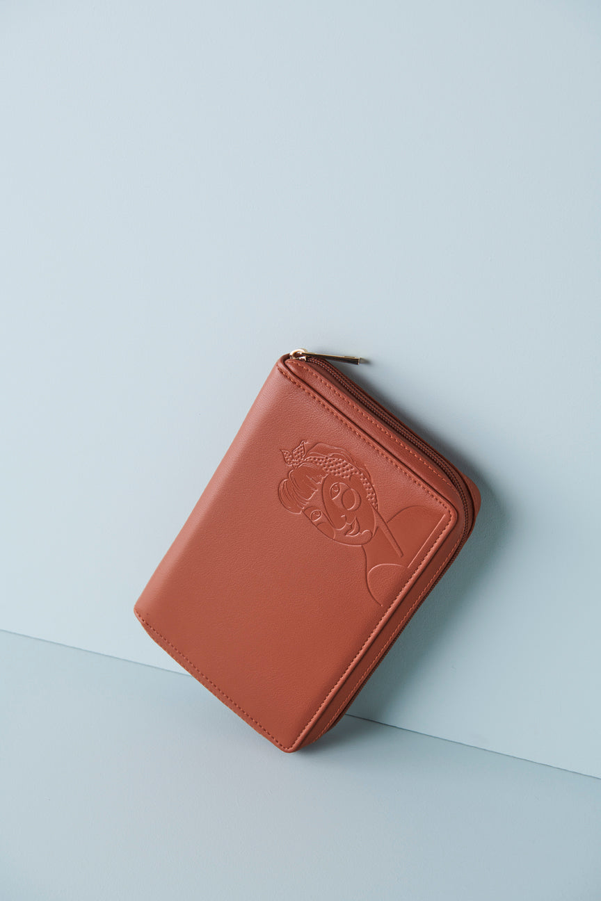 A tan jewellery wallet sits on an angle against a baby blue background. The wallet features an embossed image of a lady with a fringe and head scarf. It has a tan zip and has a gold zip pull.