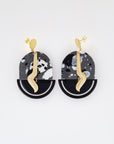 A pair of stud dangle earrings sit against a white background. They feature a black and grey acrylic arch, a black enamel D- shape piece, and a brass drop with a wave.