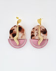 A pair of stud dangle earrings sit against a white background. They feature a brown and cream acrylic arch, a mauve enamel D- shape piece, and a brass drop with a wave.
