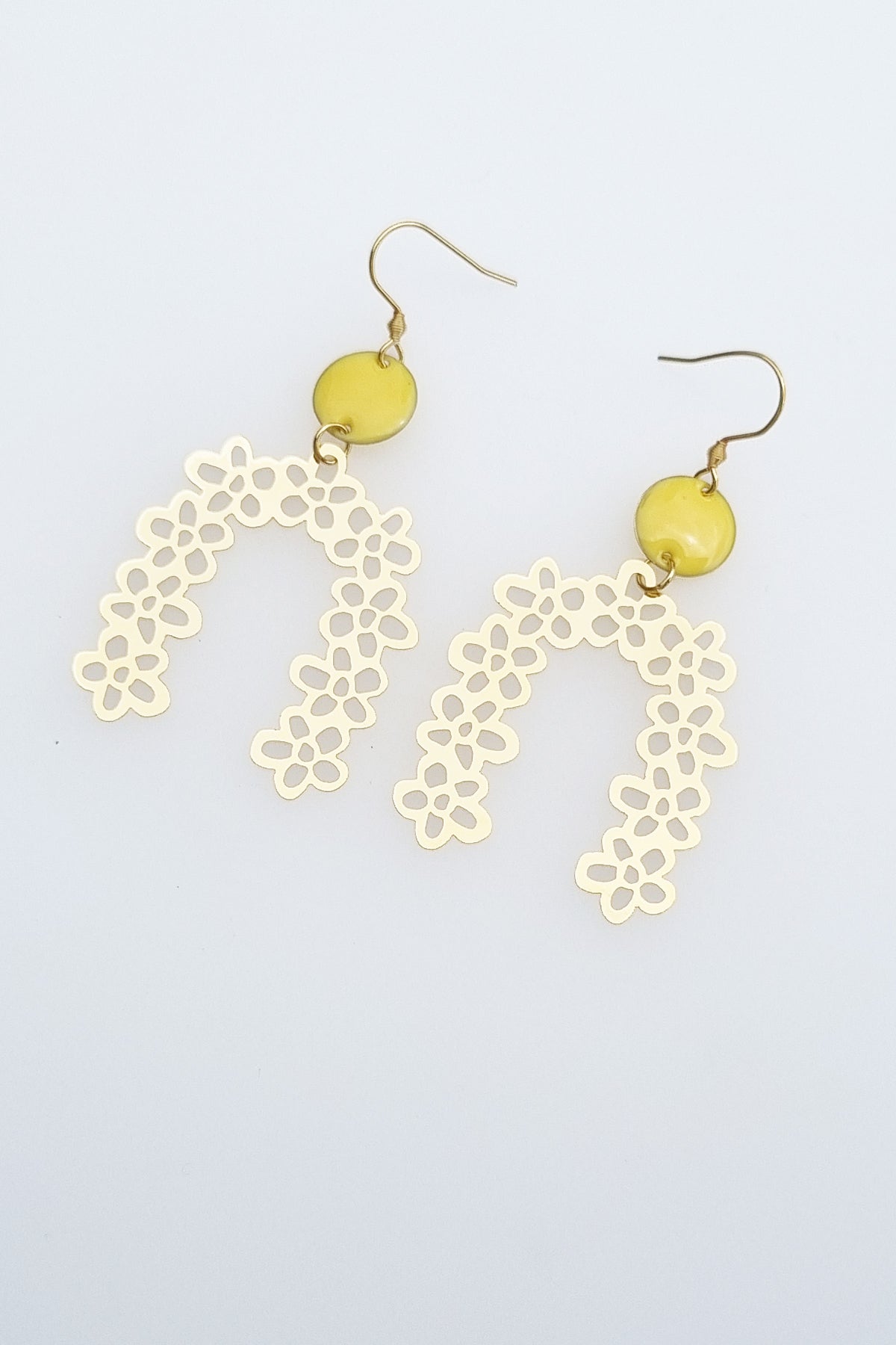 A pair of drop earrings with a hook lay against a white background. They feature a small yellow connector dot and a floral arch brass piece. 