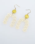 A pair of drop earrings with a hook lay against a white background. They feature a small yellow connector dot and a floral arch brass piece. 