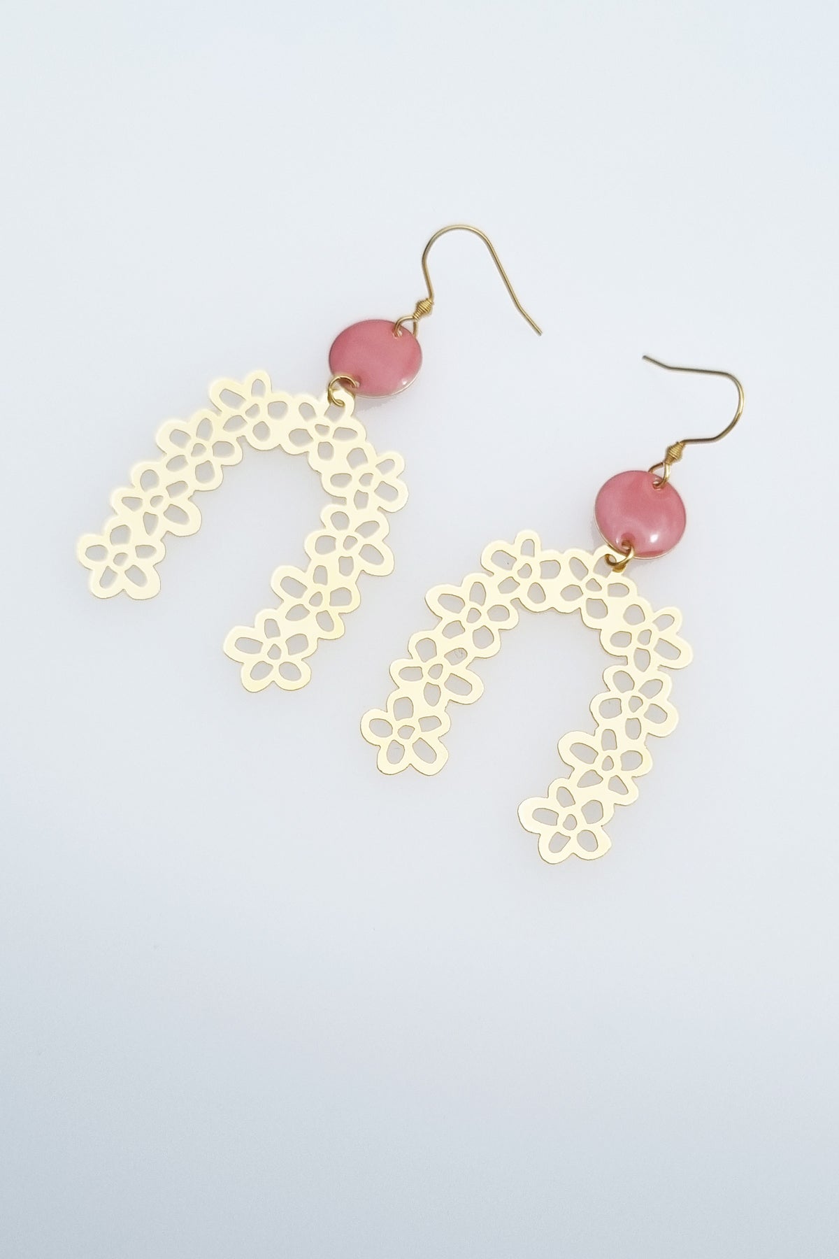 A pair of drop earrings with a hook lay against a white background. They feature a small pink connector dot and a floral arch brass piece.