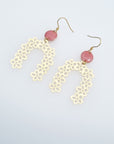 A pair of drop earrings with a hook lay against a white background. They feature a small pink connector dot and a floral arch brass piece.