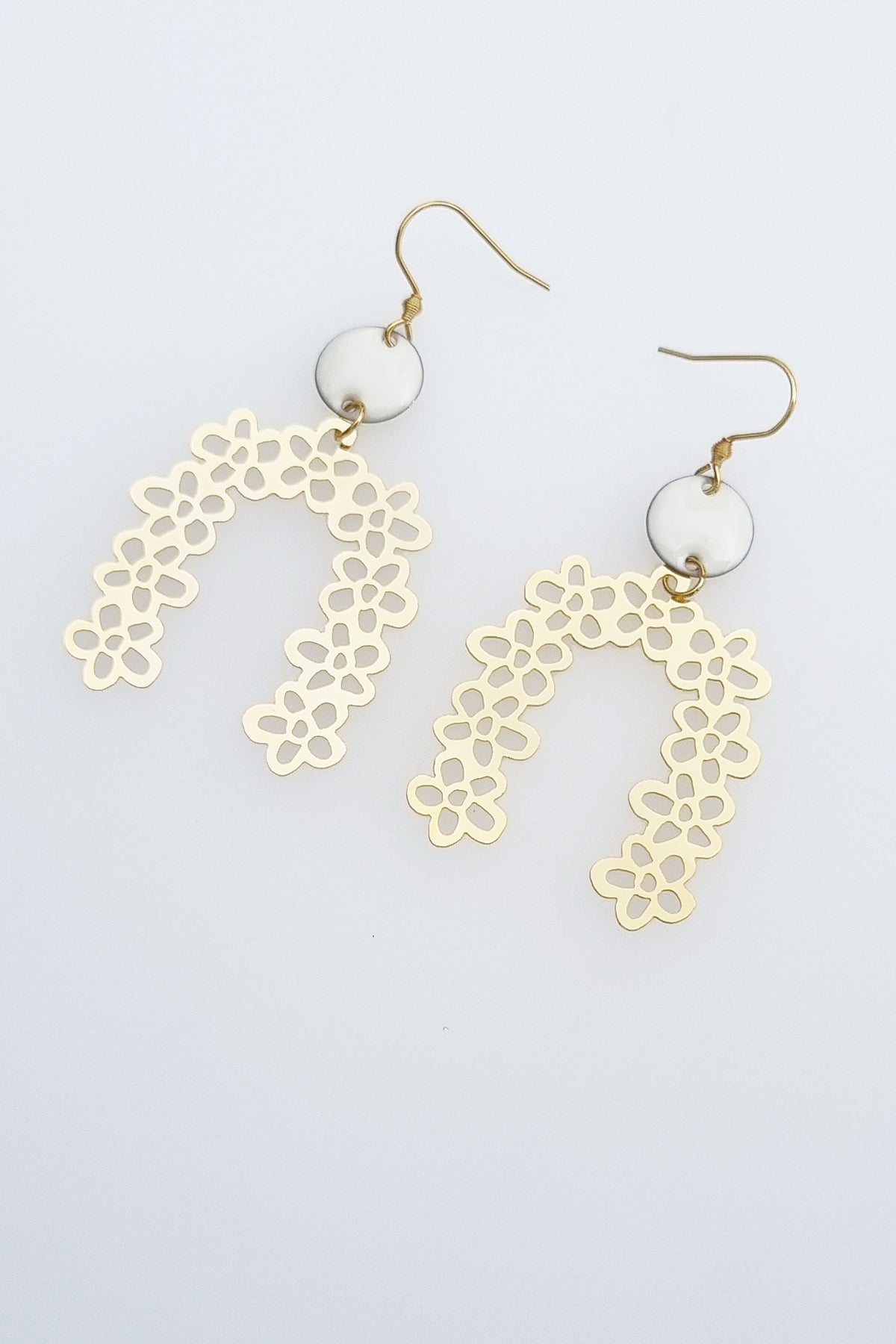 A pair of drop earrings with a hook lay against a white background. They feature a small white connector dot and a floral arch brass piece.