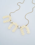 A necklace sits against a white background. It features a gold chair with three floral brass arch pieces, and two white enamel connector dots separating each arch.
