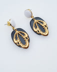 A pair of drop earrings sit against a white background. They feature a white enamel stud top with a black oval acrylic, and a leaf-like brass piece.