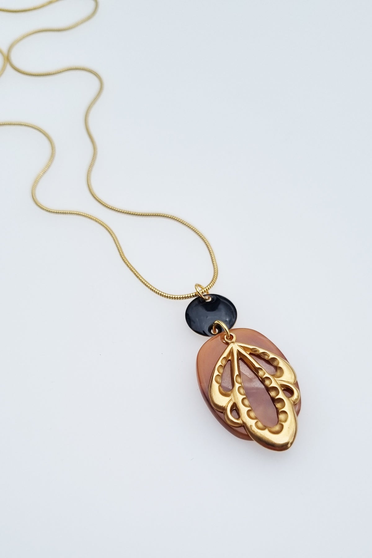 A necklace lays against a white background. It features a gold chain, a black enamel connector dot, a brown acrylic oval, and a brass leaf shape.