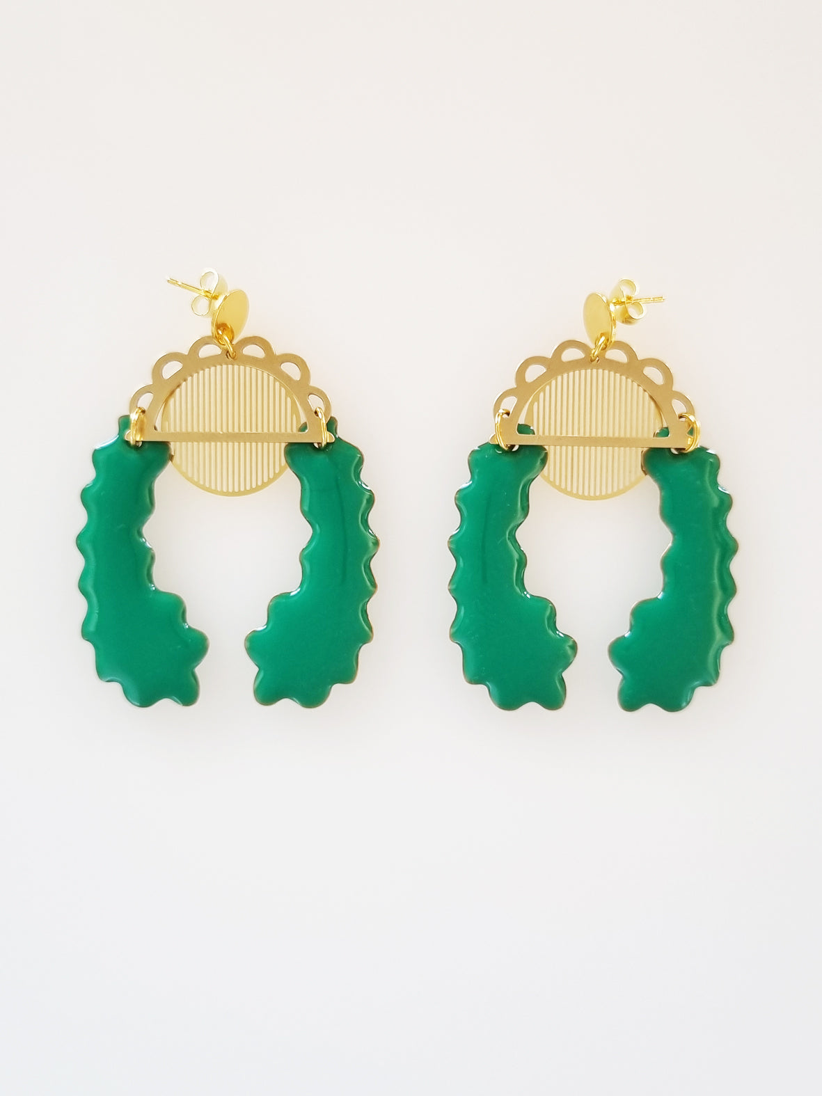 A pair of stud dangle earrings lay on a white background. Both earrings feature a semi circle piece of brass with scalloped detailing around the top curve. Behind that is a delicate circle piece of brass with thin lines cut vertically. A long emerald green piece of enamel that has wavy edges is connected to the left of the brass semi-circle with a jump ring. A second enamel piece in the same shape is mirrored and connected to the right side of the brass semi-circle.