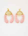 A pair of stud dangle earrings lay on a white background. Both earrings feature a semi circle piece of brass with scalloped detailing around the top curve. Behind that is a delicate circle piece of brass with thin lines cut vertically. A long pale pink piece of enamel that has wavy edges is connected to the left of the brass semi-circle with a jump ring. A second enamel piece in the same shape is mirrored and connected to the right side of the brass semi-circle.
