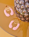 A pair of pale pink embrace earrings are pictured on a glass table. The ground is a warm yellow colour.  There is a half cut pineapple sitting on the table next two the earrings.