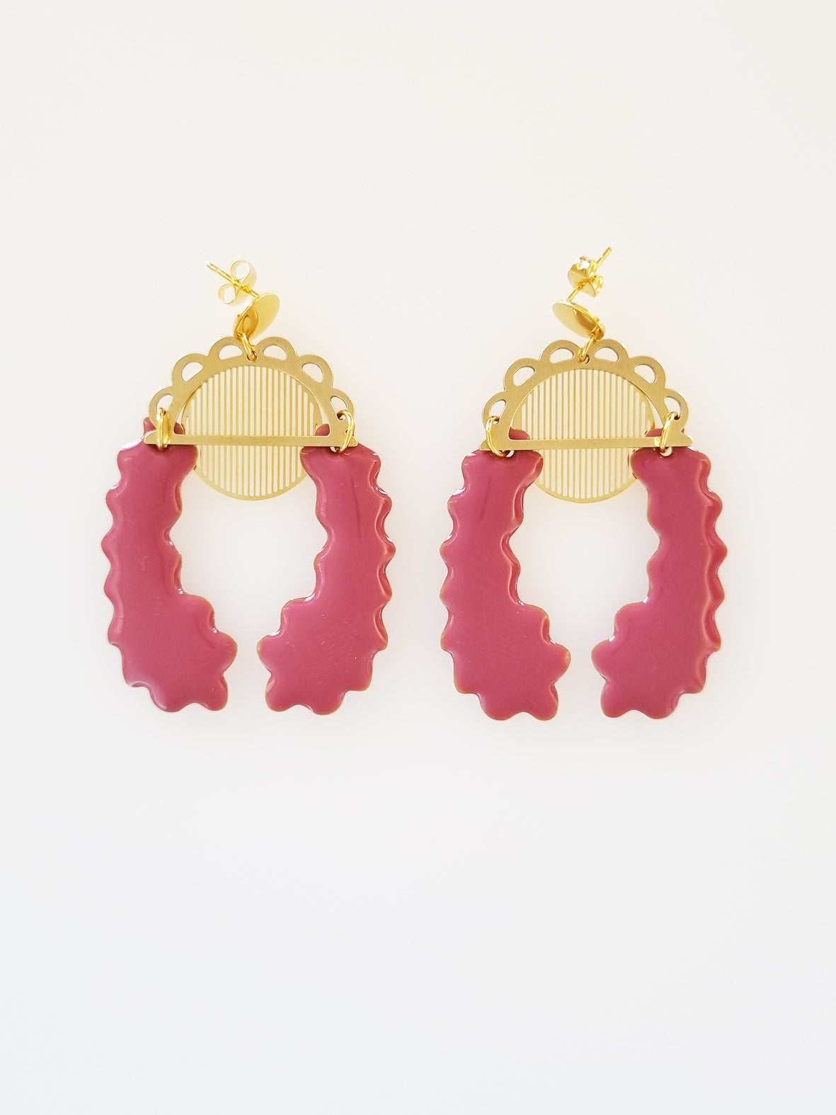 A pair of stud dangle earrings lay on a white background. Both earrings feature a semi circle piece of brass with scalloped detailing around the top curve. Behind that is a delicate circle piece of brass with thin lines cut vertically. A long plum piece of enamel that has wavy edges is connected to the left of the brass semi-circle with a jump ring. A second enamel piece in the same shape is mirrored and connected to the right side of the brass semi-circle.