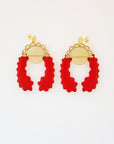 A pair of stud dangle earrings lay on a white background. Both earrings feature a semi circle piece of brass with scalloped detailing around the top curve. Behind that is a delicate circle piece of brass with thin lines cut vertically. A long red piece of enamel that has wavy edges is connected to the left of the brass semi-circle with a jump ring. A second enamel piece in the same shape is mirrored and connected to the right side of the brass semi-circle.