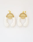A pair of stud dangle earrings lay on a white background. Both earrings feature a semi circle piece of brass with scalloped detailing around the top curve. Behind that is a delicate circle piece of brass with thin lines cut vertically. A long white piece of enamel that has wavy edges is connected to the left of the brass semi-circle with a jump ring. A second enamel piece in the same shape is mirrored and connected to the right side of the brass semi-circle.