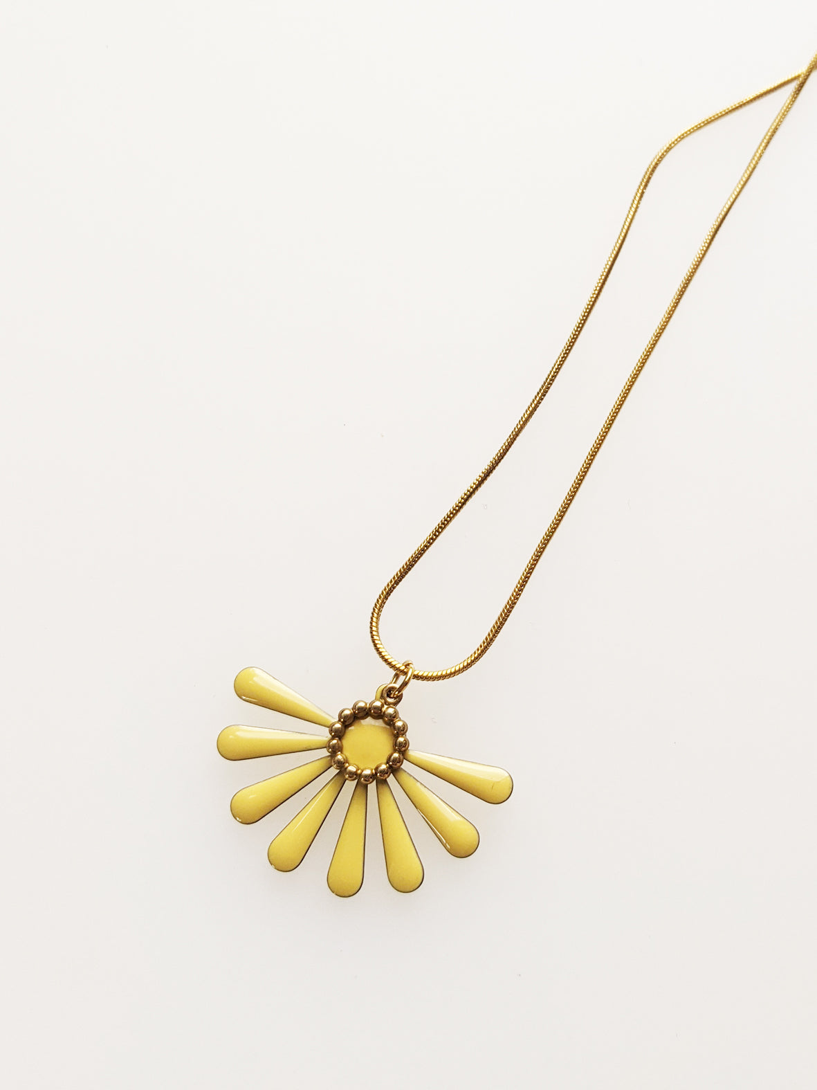 A necklace sits against a white background. It features a gold chain and a yellow coloured floral enamel shape. A tiny brass ring encircles the disk of the floral enamel piece.