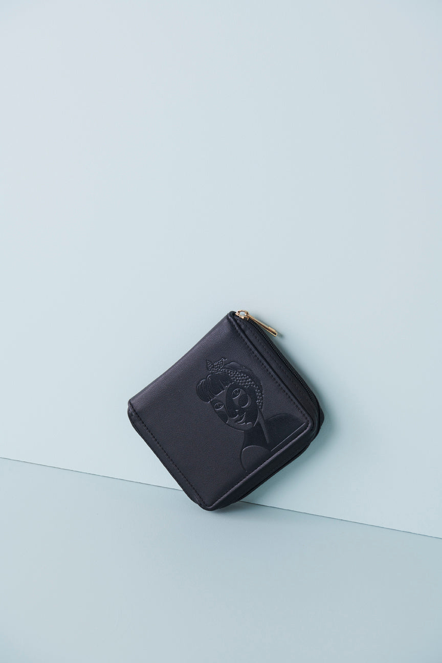A black jewellery wallet sits on an angle against a baby blue background. The wallet features an embossed image of a lady with a fringe and head scarf. It has a black zip and has a gold zip pull.