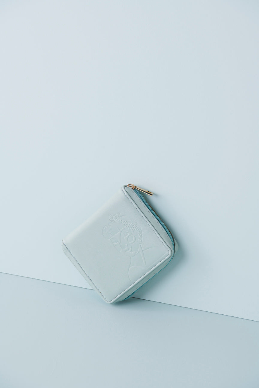 A duckegg jewellery wallet sits on an angle against a baby blue background. The wallet features an embossed image of a lady with a fringe and head scarf. It has a duckegg zip and has a gold zip pull.