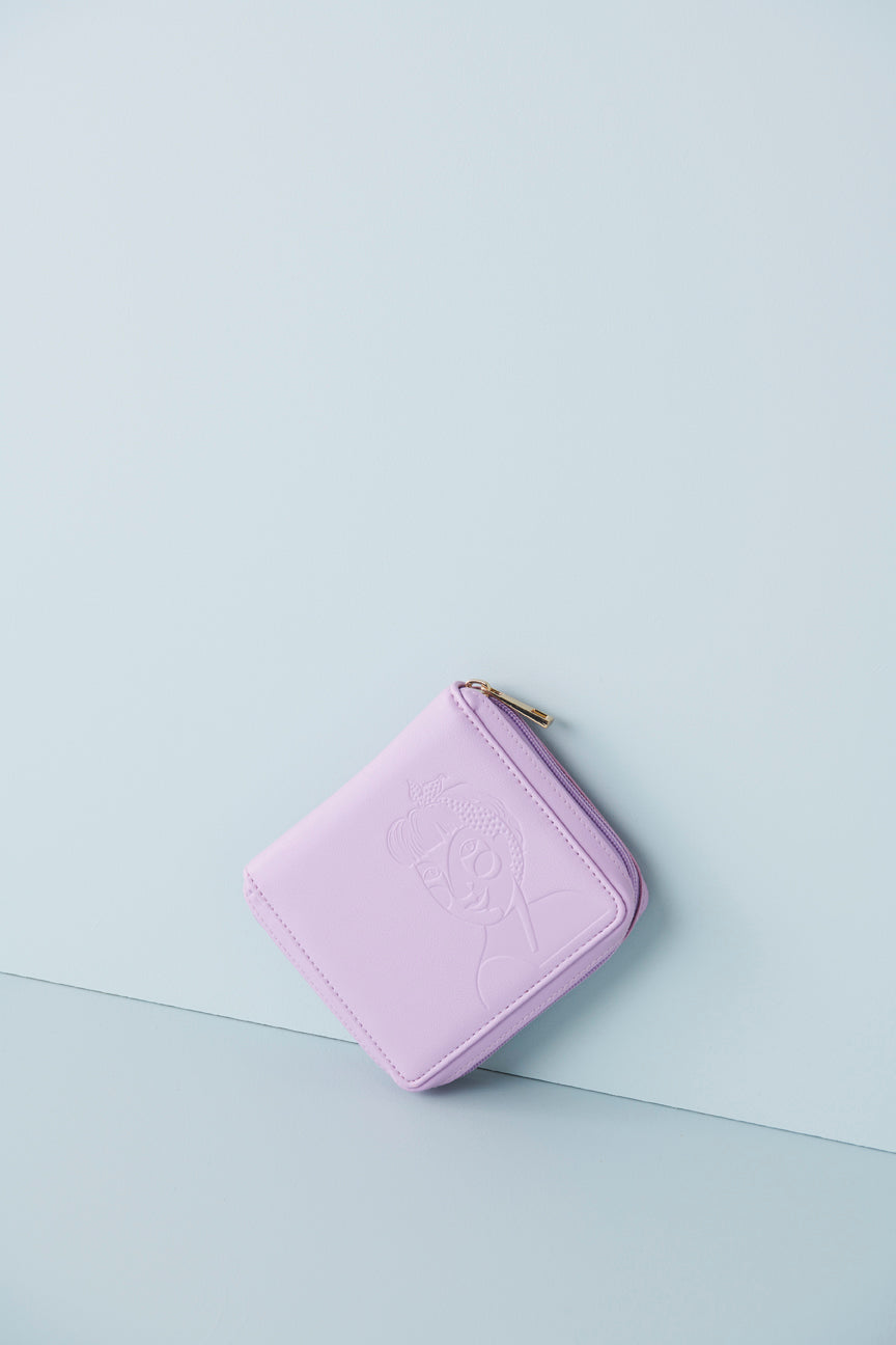 A lilac jewellery wallet sits on an angle against a baby blue background. The wallet features an embossed image of a lady with a fringe and head scarf. It has a lilac zip and has a gold zip pull.