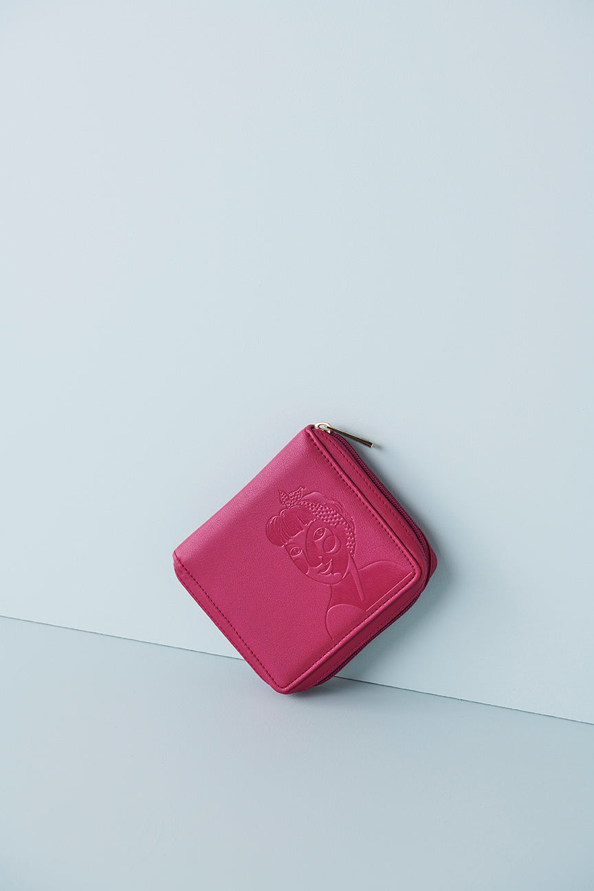 A magenta jewellery wallet sits on an angle against a baby blue background. The wallet features an embossed image of a lady with a fringe and head scarf. It has a magenta zip and has a gold zip pull.