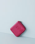 A magenta jewellery wallet sits on an angle against a baby blue background. The wallet features an embossed image of a lady with a fringe and head scarf. It has a magenta zip and has a gold zip pull.