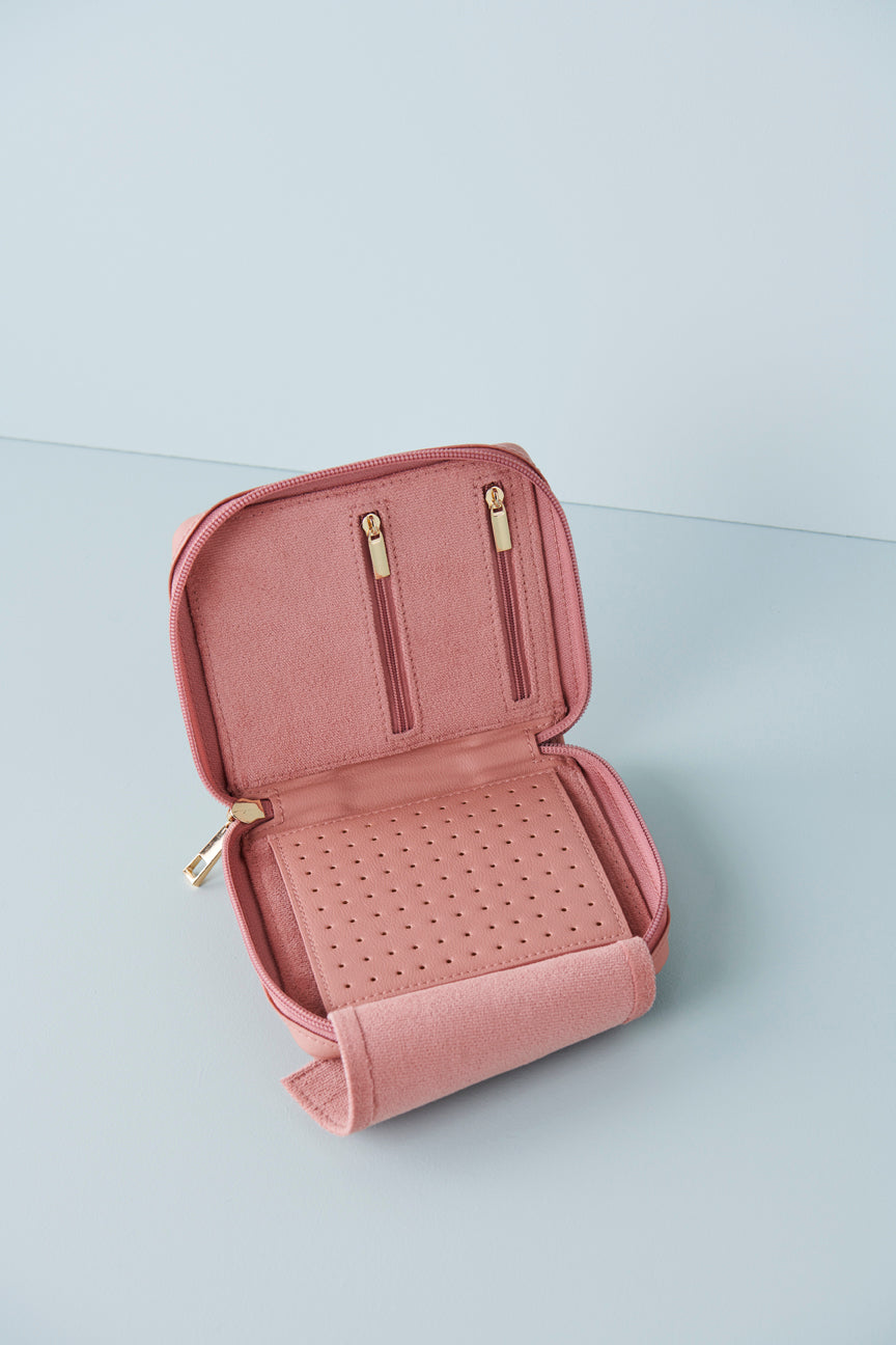 A pink wallet sits open against a baby blue background. The wallet inner is pink, the top section features two zip pockets with gold zip pulls. The bottom section features a felt flap that opens to reveal a perforated pu section for earrings.