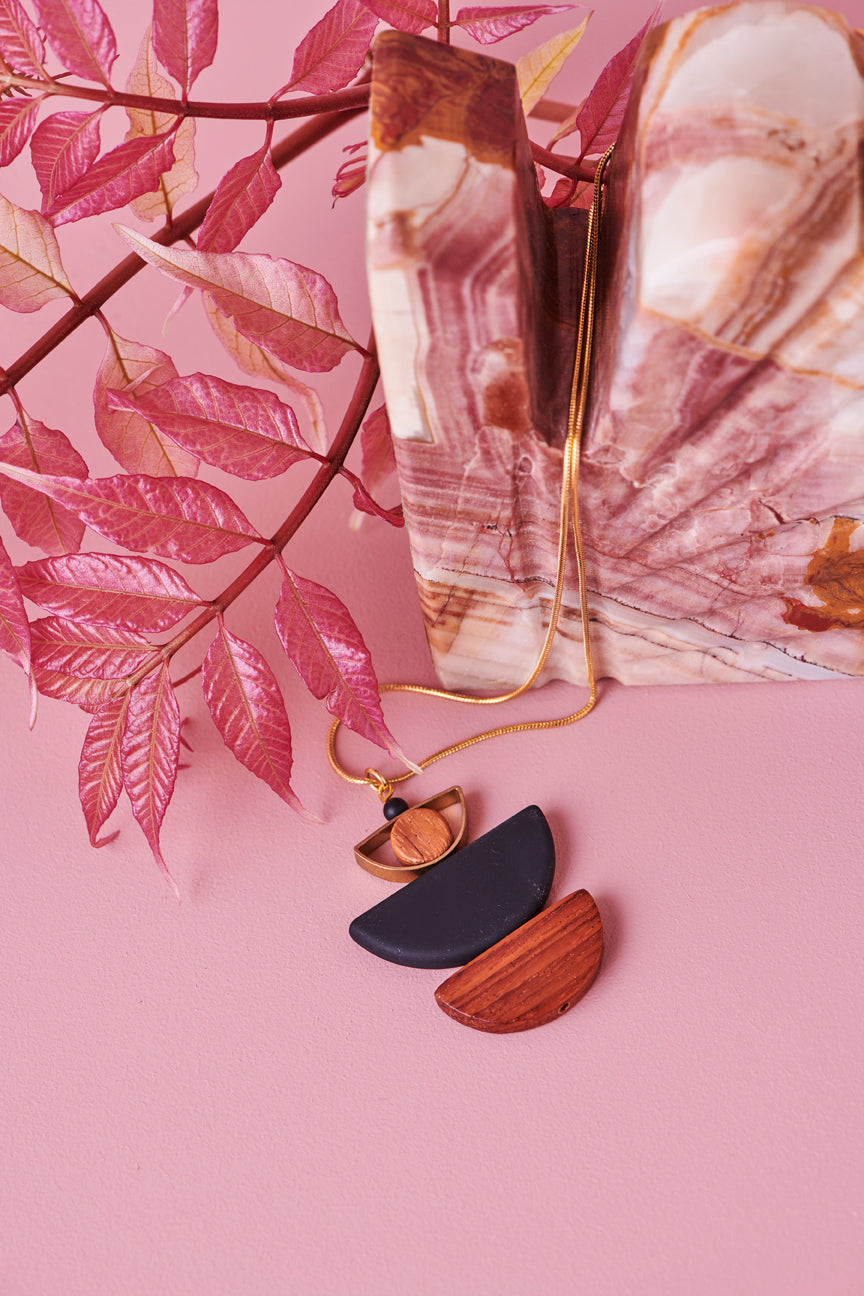 A black Marcel necklace sits styled against a pink background. The necklace chain is draped over a marble stone with pink and brown colours through it, and a branch with pink leaves sits behind the stone.